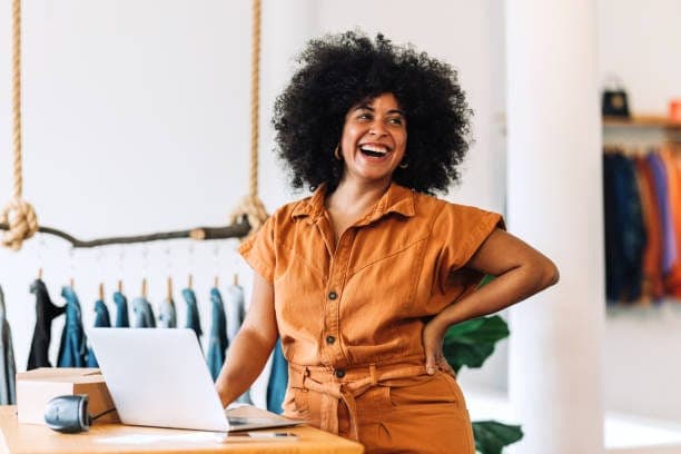 A shop owner laughing at her counter, with clothes on the racks behind her